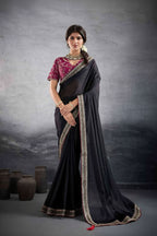 A woman modeling a traditional Indian saree in a studio setting, shown in two side-by-side views. The saree is black with a metallic gold border, paired with a rich magenta pink embroidered blouse.