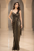 A woman modeling a floor-length draped evening gown in a grand arched hall. The image shows the same dress twice side by side, once in deep navy blue and once in metallic taupe-brown, both with one-shoulder draping and embellished bodice details.