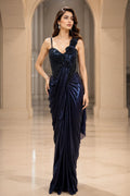Two women modeling floor-length evening gowns in an arched indoor hallway. The left gown is dark navy blue with a ruched bodice and sheer draped skirt.