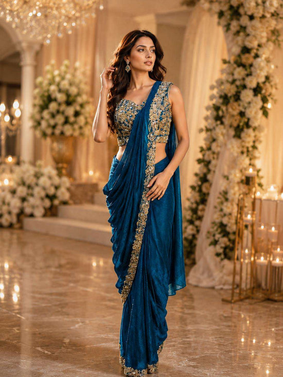 A woman modeling a royal blue saree with intricate gold embroidery on the blouse and border, set in an elegant indoor venue with warm lighting and floral decorations.