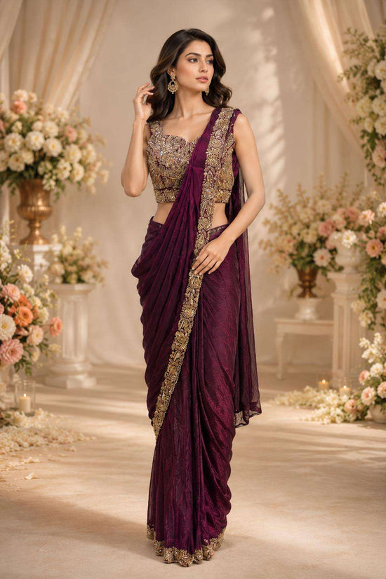 A woman models a deep purple saree with gold embroidery and a matching embroidered blouse in a decorated indoor setting with floral arrangements and draped curtains.