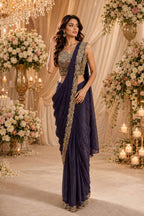 A woman models a dark purple saree with gold embroidery, paired with a matching embroidered blouse, set against a decorated event backdrop with flowers and candles.