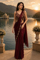 A woman stands by a lake at sunset wearing a deep maroon saree with gold embroidery and a matching embellished sleeveless blouse. Mountains and water are visible in the background.