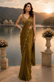 A woman stands outdoors by a lake at sunset wearing an olive green saree with a heavily embroidered gold blouse. Mountains, water, and a domed building are visible in the background.