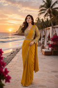 A woman stands on a beach walkway at sunset wearing a flowing mustard yellow sari with heavily embellished gold sleeves and a matching embellished waistband. Palm trees, ocean, and lounge seating are visible in the background.
