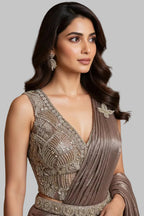 A woman models a floor-length metallic taupe gown with a deep V-neckline, heavily embellished bodice, and draped side panel in an elegant indoor hallway with chandeliers and polished floors.