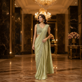 A woman models a light sage green saree with intricate silver embroidery on the border and a matching embellished sleeveless blouse, standing in a luxurious hallway with marble columns and chandeliers.