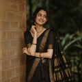 A woman wearing a black saree with gold zari patterns leans against a brick wall outdoors, with a blurred green foliage background.