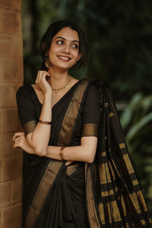 A woman wearing a black saree with gold zari patterns leans against a brick wall outdoors, with a blurred green foliage background.