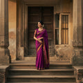 Woman in a purple saree standing in front of a rustic building.
