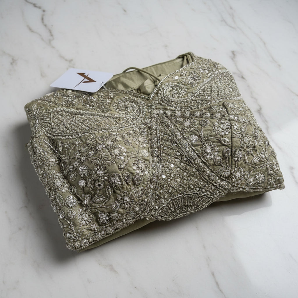 An olive green embroidered ethnic garment, likely a kurta or suit piece, featuring intricate silver sequin and bead work, folded on a matching olive green fabric background with a blue price tag attached.