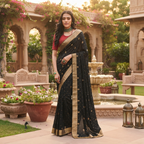 A woman stands in an outdoor courtyard wearing a black saree with gold motifs and a red blouse. The background features stone arches, climbing plants, and a fountain.
