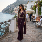 A woman wearing a deep purple sleeveless maxi dress with gold embroidery and sheer sleeves, standing on a stone walkway overlooking the sea and a coastal town.
