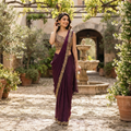 A woman stands in a sunlit courtyard wearing a deep purple saree with gold embroidery and a matching embroidered blouse. The setting features stone paving, potted plants, and a fountain in the background.