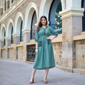 A woman modeling a teal green A-line kurta dress with black embroidered details on the pockets and neckline, standing outdoors in front of a stone building. The brand logo 'KADLEE' is visible in the top right corner.