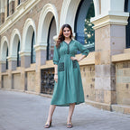 A woman modeling a teal green A-line kurta dress with black embroidered details on the pockets and neckline, standing outdoors in front of a stone building. The brand logo 'KADLEE' is visible in the top right corner.