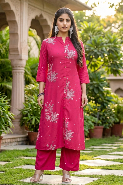 A woman stands outdoors in a garden courtyard wearing a bright pink traditional kurta set with white floral embroidery and matching pants.