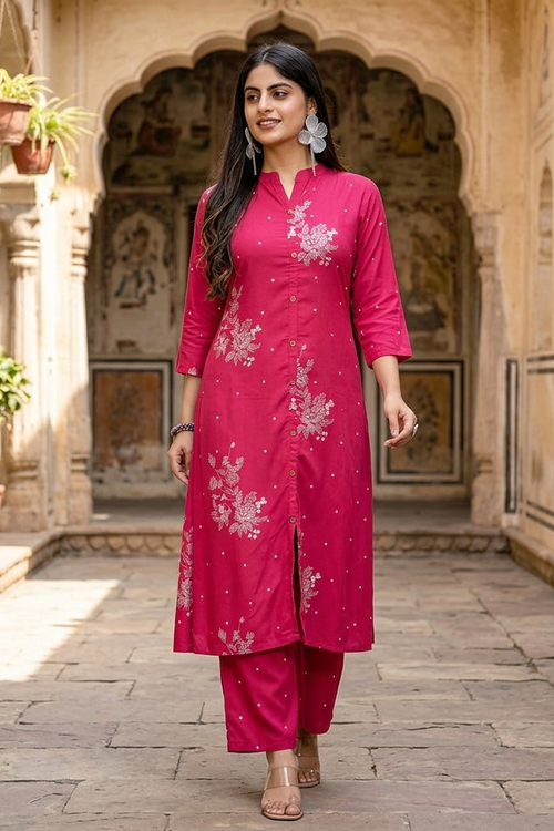 A woman stands in a courtyard wearing a bright pink traditional kurta set with white floral embroidery and small metallic embellishments. The setting features stone arches, potted plants, and warm architectural details.