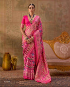A woman modeling a bright pink saree with intricate gold and multicolor floral patterns, shown in a split-screen presentation against a vintage-style beige background with a carved sofa and decorative vases.