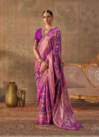 A woman models a purple and pink patterned saree with a matching purple blouse in an ornate room with a patterned wall, gold sofa, and decorative vases. The brand name 'KRESHVA' is visible at the top.