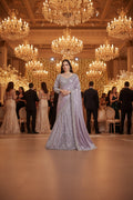 A woman modeling a lavender purple embroidered traditional Indian gown with a matching dupatta, standing in front of a patterned beige and gold wall backdrop.