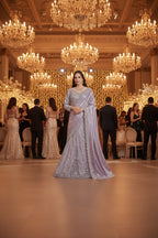 A woman modeling a lavender purple embroidered traditional Indian gown with a matching dupatta, standing in front of a patterned beige and gold wall backdrop.