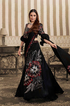 A woman models a long black embroidered gown with red, white, and silver floral patterns in an ornate indoor setting with striped wallpaper and a patterned rug.