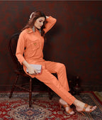 A woman models a matching burnt orange two-piece jumpsuit set, consisting of a long-sleeve button-up shirt with embroidered details on the chest and matching trousers. She is seated on a dark wooden chair against a dark textured wall, holding a small white clutch.