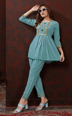A woman models a light blue-green two-piece kurta set with gold embroidery and tassel trim, standing against a dark brown textured wall.