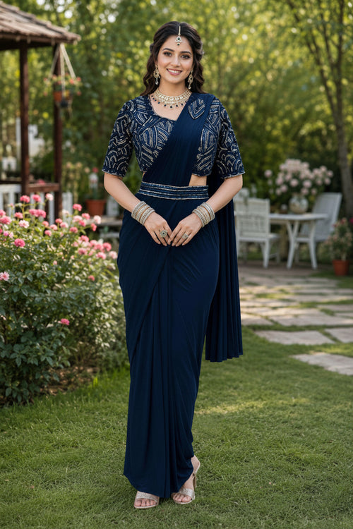 A woman stands outdoors on a grassy garden path wearing a dark navy blue traditional outfit with an embroidered blouse and draped skirt. The background shows green foliage, flowers, and patio furniture.