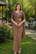 A woman stands outdoors in a garden wearing a brown traditional sari with a heavily embroidered dark brown blouse. The setting features green grass, trees, and patio furniture in the background.