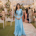 Woman in a light blue traditional outfit at an event with floral decorations and people in the background.