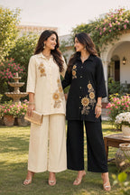 Two women standing outdoors in a garden setting, wearing matching embroidered kurta-and-pant sets. One outfit is cream with gold floral embroidery, and the other is black with similar gold embroidery.