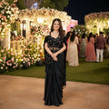 Woman in a black saree with floral patterns at an event with decorative lights and people in the background.