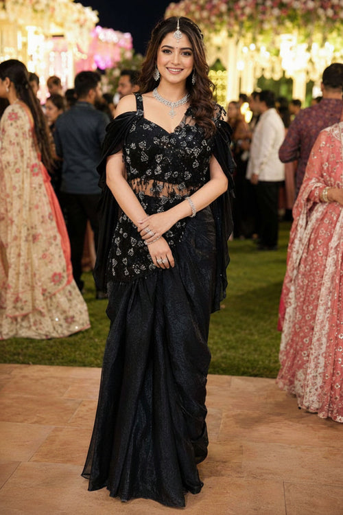 Woman in a black saree with floral patterns at an event with decorative lights and people in the background.