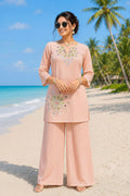 Woman in peach embroidered outfit standing on a beach with palm trees in the background