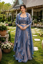 Woman in a blue traditional outfit standing outdoors with greenery and a house in the background