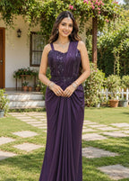 A woman stands outdoors in a garden wearing a floor-length sleeveless gown in deep purple with embroidered detailing on the bodice and draped fabric at the skirt. The background shows green grass, potted plants, and a house entrance.