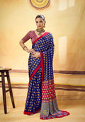 A woman models a blue and red saree with gold and multicolor patterns, standing in front of a warm beige backdrop with a small wooden stool on the left.