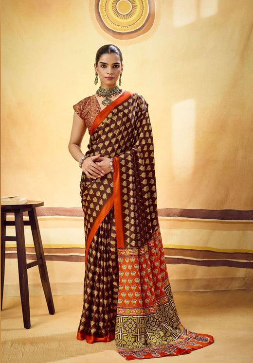 A woman models a brown and orange saree with intricate gold and red patterns, standing in front of a beige studio backdrop with a wooden stool on the left.
