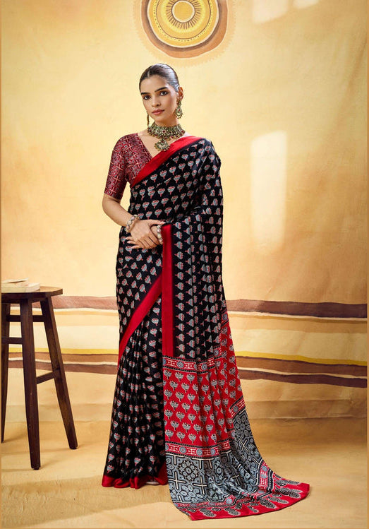 A woman models a black and red saree with intricate geometric patterns, standing in front of a warm beige studio backdrop with a wooden stool on the left.