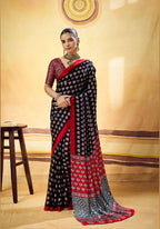 A woman models a black and red saree with intricate geometric patterns, standing in front of a warm beige studio backdrop with a wooden stool on the left.