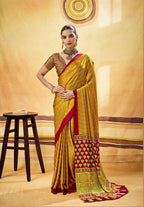 A woman models a yellow and red saree with intricate patterns and a gold embroidered blouse, standing in front of a warm beige textured wall with a wooden stool to the side.