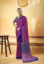 A woman models a purple and blue patterned saree with a matching blue blouse in a warm beige studio setting. The saree features intricate geometric and floral motifs in purple and blue, with a detailed border.
