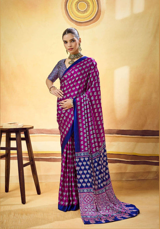 A woman models a purple and blue patterned saree with a matching blue blouse in a warm beige studio setting. The saree features intricate geometric and floral motifs in purple and blue, with a detailed border.