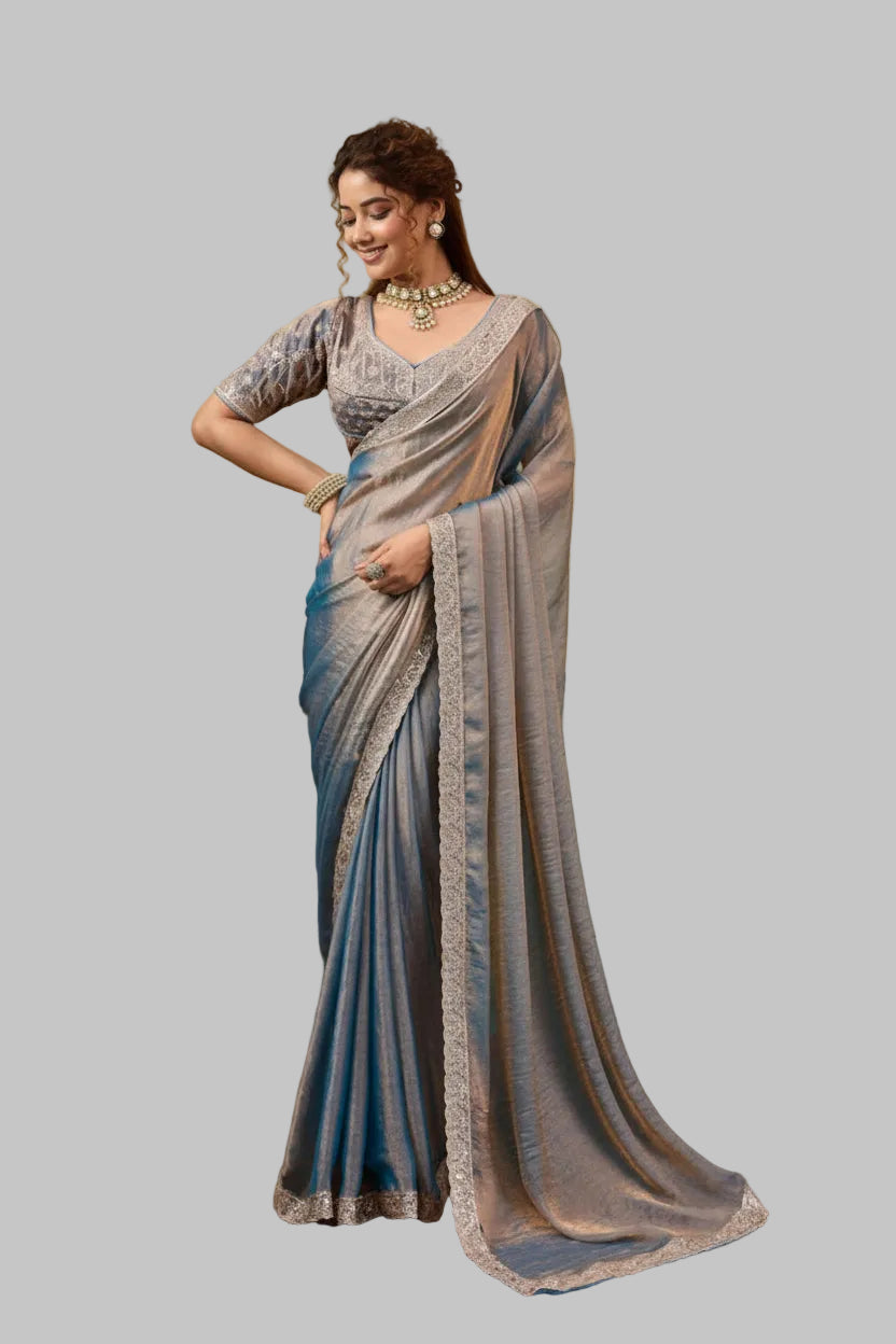 A woman models a blue and gray saree with a matching printed blouse in front of a tropical-themed wall mural. The saree has a gray-blue body, a blue border, and a sheer dupatta draped over her shoulder.