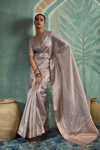 A woman models a light lavender-gray silk saree with subtle gold floral embroidery and a matching embroidered border, posed against a textured teal wall with decorative clay pots.