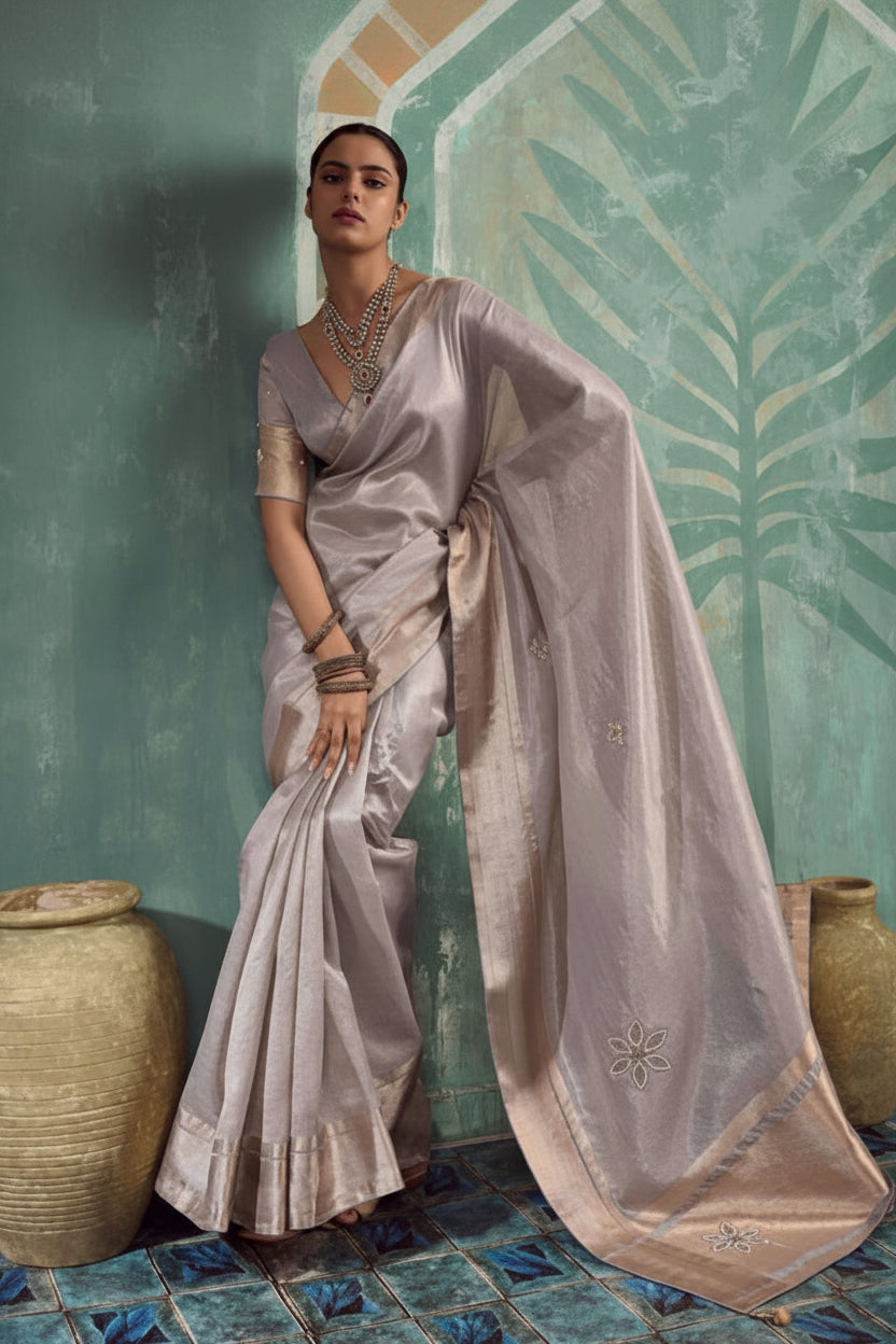 A woman models a light lavender-gray silk saree with subtle gold floral embroidery and a matching embroidered border, posed against a textured teal wall with decorative clay pots.