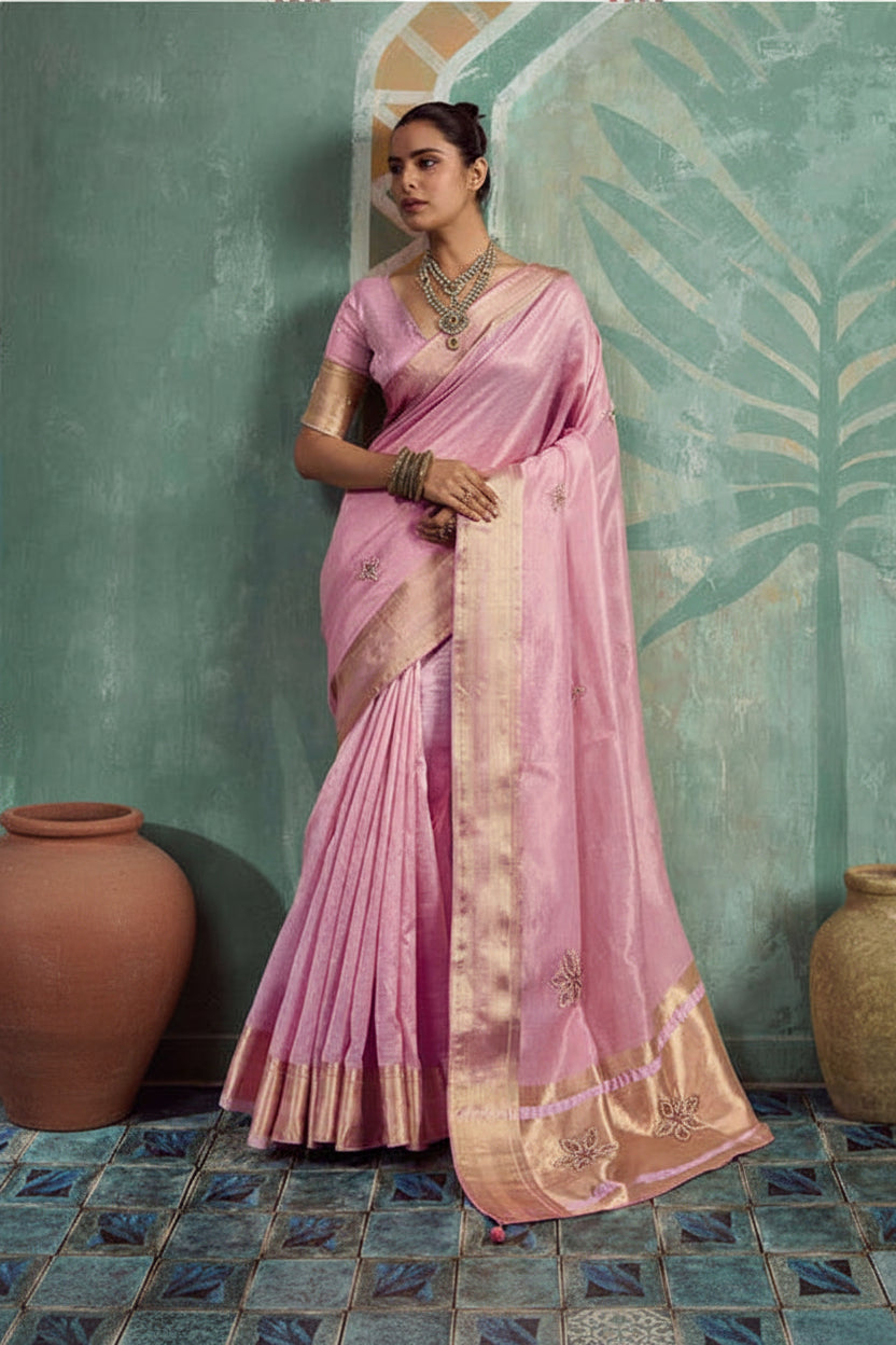 A woman models a light pink silk saree with a wide gold border and matching gold embroidered blouse. She stands against a textured teal wall with painted palm leaves, flanked by two decorative clay pots.