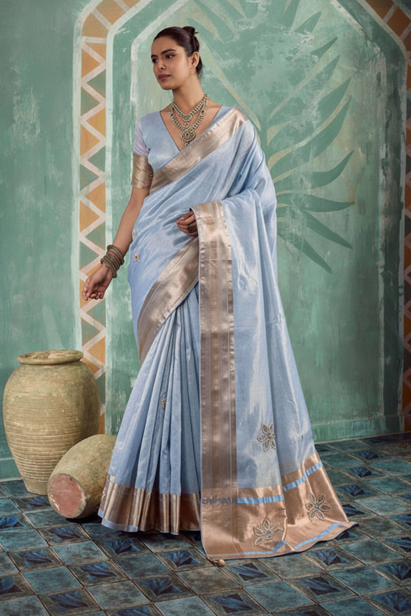 A woman models a light blue saree with a metallic beige border and floral embroidery, posed in front of a decorative teal archway with clay pots on the floor.